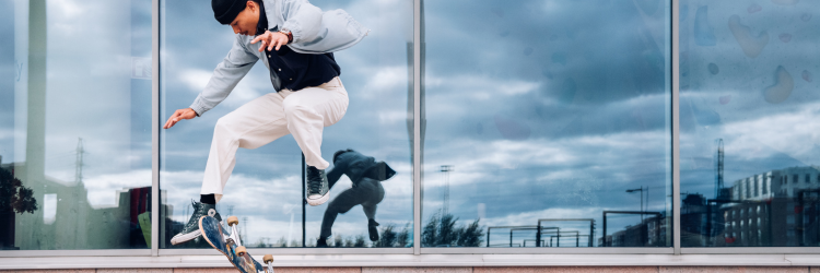 Man with a skateboard in front of a glass building. The man is preforming a stunt on his skateboard and his reflection can be seen in the glass behind.