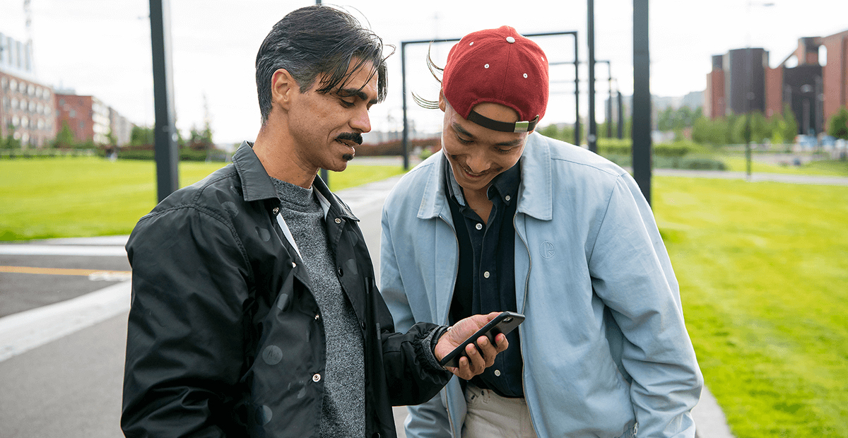 Två män tittar på en smartphone tillsammans. Den ena mannen har mörkt hår, en mustasch, och bär en svart jacka över en grå tröja. Den andra mannen, som bär en röd keps och en ljusblå jacka, ser ut att peka på något på skärmen. 