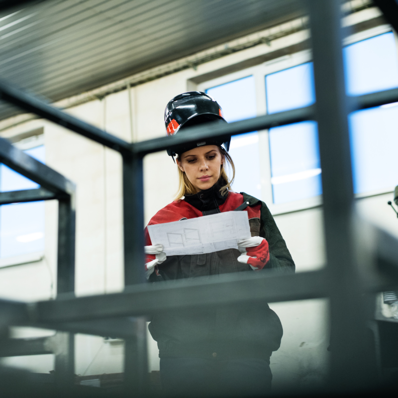 en kvinna står i svarta och röda arbetskläder i en industriell lokal. hon tittar på en ritning