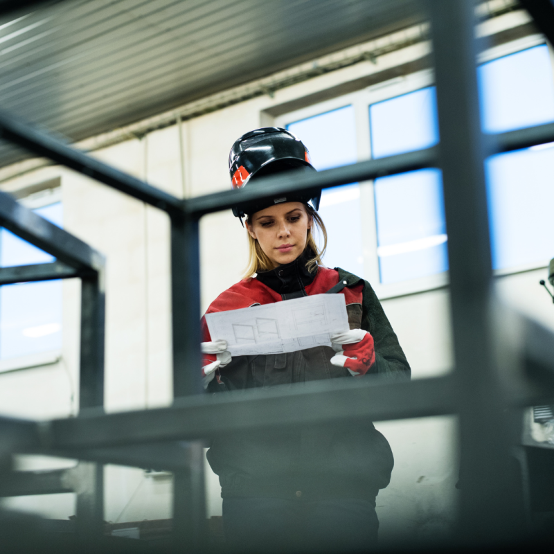 a woman stands in black and red work clothes in an industrial premises. she is looking at a drawing