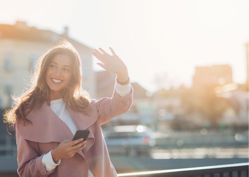 Decoartive image of a person standing in a sunny harbor with her phone in her hand, she is waving and smiling