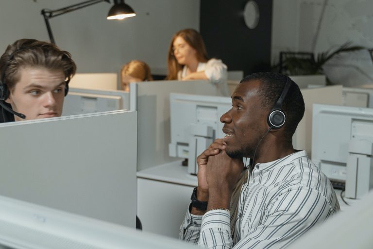 miljöbild från ett kontaktcenter med kundserviceagenter som arbetar. två män sitter med hörlurar i samtal och två kvinnor syns i bakgrunden där de samarbetar framför en datorskärm