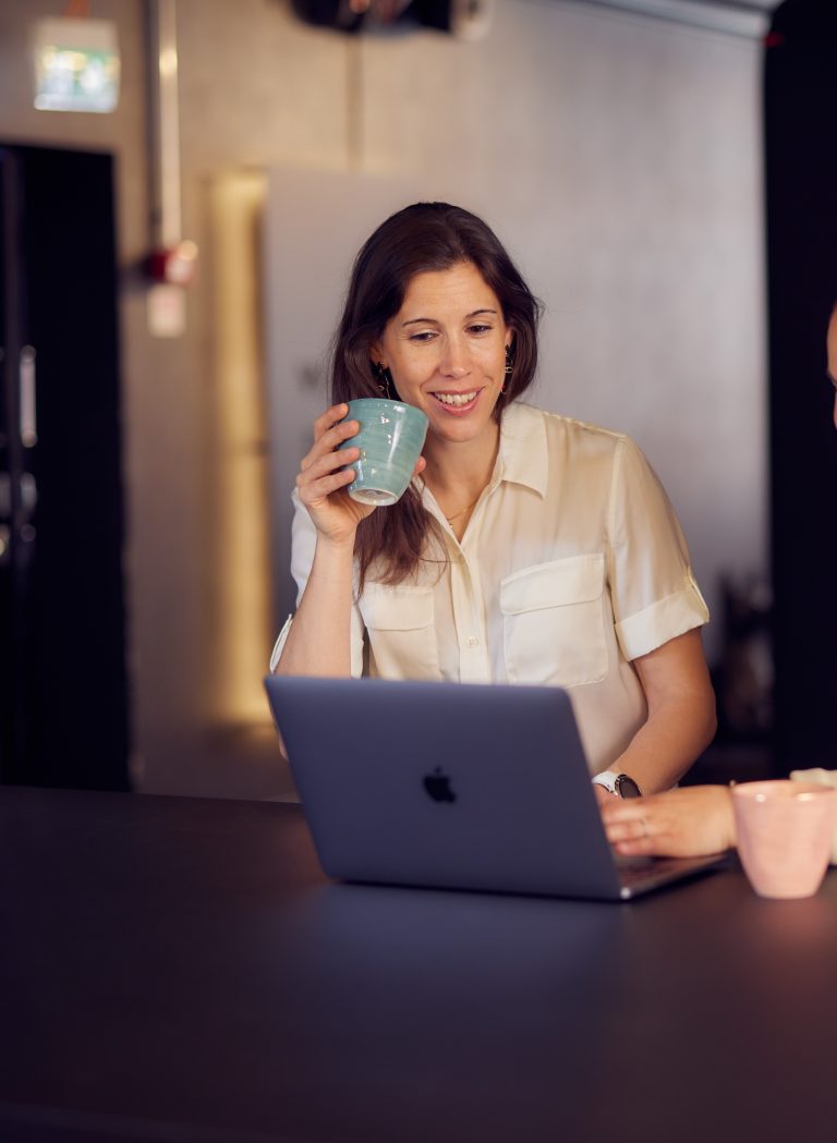 Bild på en kvinna som sitter framför en laptop och dricker kaffe.