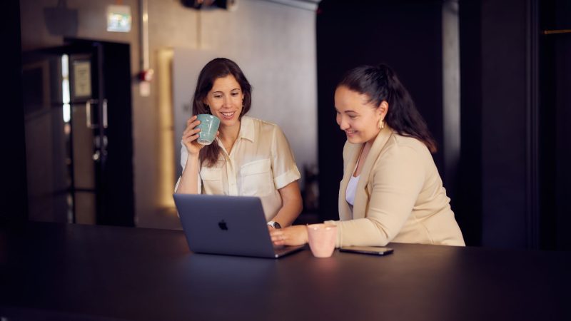 Två kvinnor vid ett bord med en bärbar dator och kaffe.