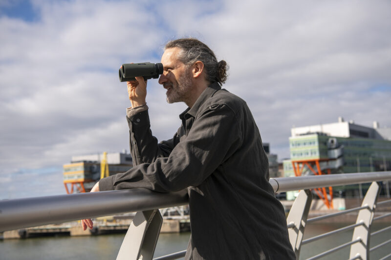Dekorativ bild av en man som står med en kikare på en bro och tittar ut över en stad.