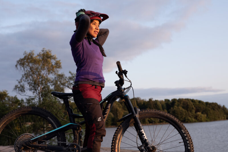 Dekorativ bild av en person som står med sin cykel, hen har en hjälm på huvudet och tittar ut över ett vatten