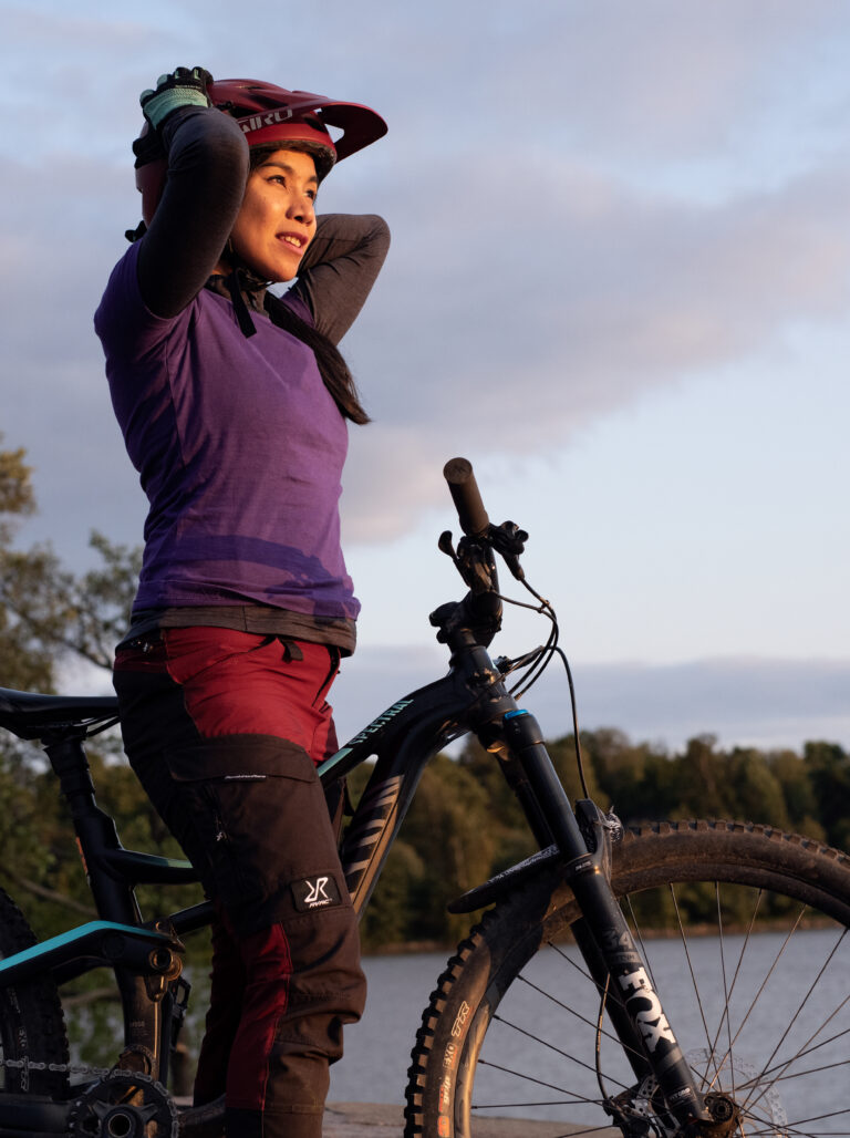 Dekorativ bild av en person som står med sin cykel, hen har en hjälm på huvudet och tittar ut över ett vatten